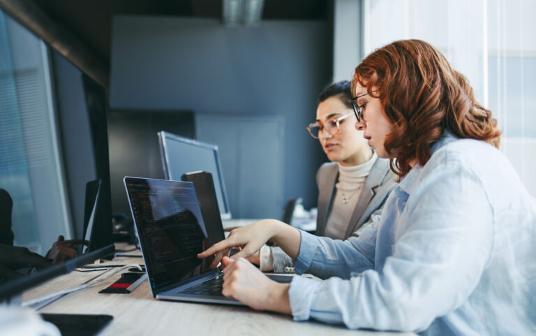 A diverse group of professionals discuss and collaborate in a modern office. They work on laptops, focusing on software design and programming. With teamwork and expertise, they strive to create innovative solutions.