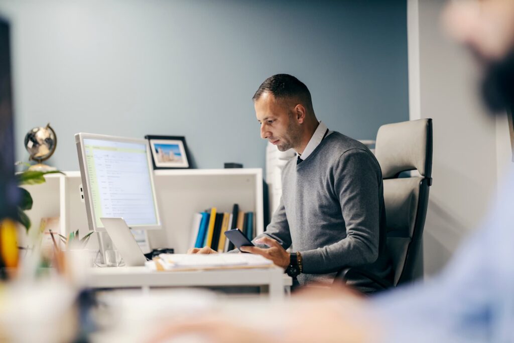 Man sitting at desk holding phone and looking at laptop